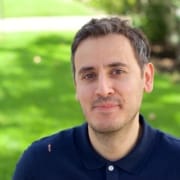 Person seated on grass wearing a navy blue polo shirt with small embroidered motifs, bright green lawn and trees in the background.