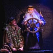 Two actors appear on a s hip with one caught up in netting and seaweed and the other portraying a ship captain with a long white beard who is gripping a ship's wheel