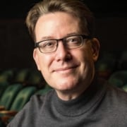 Person wearing a dark turtleneck sweater seated in a theater with green upholstered seats in the background.