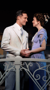 Two people stand close together on a decorative white railing, holding hands. One is wearing a light-colored pinstripe suit with a tie, and the other is dressed in a lavender gown with floral embellishments and flowers in the hair. The background suggests an evening outdoor setting.