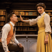 Two people are in a library setting with shelves of books behind them. One person, dressed in a rust-colored vest and striped trousers, is kneeling, while the other, wearing a long yellow dress with a white blouse, stands with an arm extended forward, palm facing outward.
