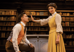 Two people are in a library setting with shelves of books behind them. One person, dressed in a rust-colored vest and striped trousers, is kneeling, while the other, wearing a long yellow dress with a white blouse, stands with an arm extended forward, palm facing outward.