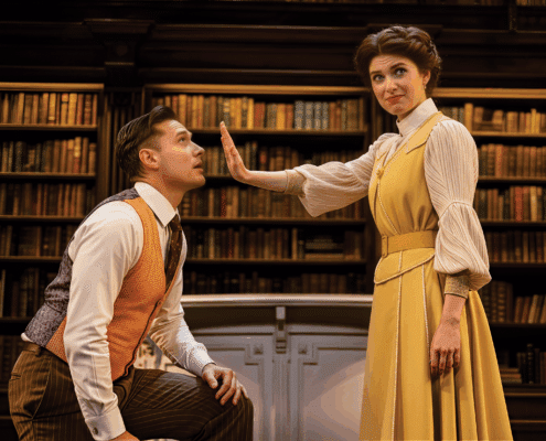 Two people are in a library setting with shelves of books behind them. One person, dressed in a rust-colored vest and striped trousers, is kneeling, while the other, wearing a long yellow dress with a white blouse, stands with an arm extended forward, palm facing outward.