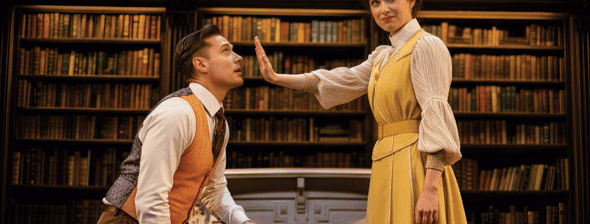 Two people are in a library setting with shelves of books behind them. One person, dressed in a rust-colored vest and striped trousers, is kneeling, while the other, wearing a long yellow dress with a white blouse, stands with an arm extended forward, palm facing outward.