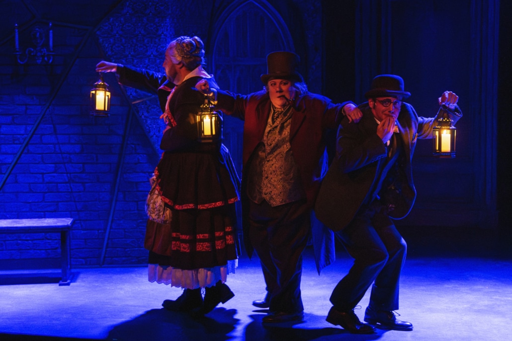 Three actors in Victorian-style costumes stand on a dimly lit stage holding lanterns that cast a warm glow. The figures are positioned in a line, leaning in different directions as if sneaking or searching. The background features a dark brick wall, a wooden bench, and an arched door, with deep blue lighting creating a mysterious, dramatic atmosphere.