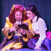Two actors in ornate Victorian-style costumes sit closely together on stage, reading from an open book. One wears a bright yellow and purple layered gown with ruffled details, while the other wears a white lace blouse paired with a deep purple skirt accented by a pink sash. The background features paneled walls in rich blue tones, creating an intimate and dramatic setting.