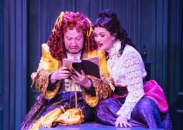 Two actors in ornate Victorian-style costumes sit closely together on stage, reading from an open book. One wears a bright yellow and purple layered gown with ruffled details, while the other wears a white lace blouse paired with a deep purple skirt accented by a pink sash. The background features paneled walls in rich blue tones, creating an intimate and dramatic setting.
