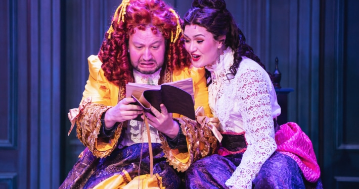 Two actors in ornate Victorian-style costumes sit closely together on stage, reading from an open book. One wears a bright yellow and purple layered gown with ruffled details, while the other wears a white lace blouse paired with a deep purple skirt accented by a pink sash. The background features paneled walls in rich blue tones, creating an intimate and dramatic setting.