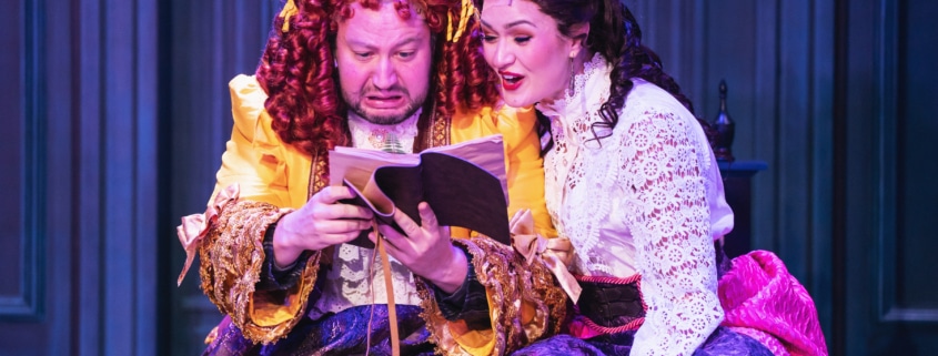 Two actors in ornate Victorian-style costumes sit closely together on stage, reading from an open book. One wears a bright yellow and purple layered gown with ruffled details, while the other wears a white lace blouse paired with a deep purple skirt accented by a pink sash. The background features paneled walls in rich blue tones, creating an intimate and dramatic setting.