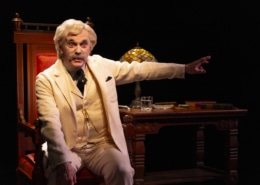 A person dressed in a white three-piece suit sits on an ornate wooden chair next to a matching desk with a stained-glass lamp, books, and a glass of water, extending one arm outward under dramatic stage lighting.