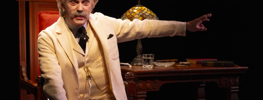 A person dressed in a white three-piece suit sits on an ornate wooden chair next to a matching desk with a stained-glass lamp, books, and a glass of water, extending one arm outward under dramatic stage lighting.