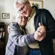 Elderly couple dancing together in a cozy living room with framed artwork and a bookshelf in the background.