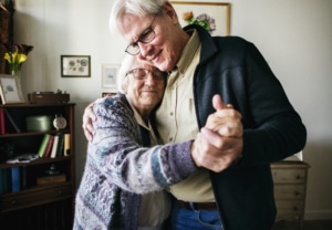 Elderly couple dancing together in a cozy living room with framed artwork and a bookshelf in the background.