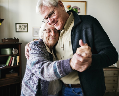 Elderly couple dancing together in a cozy living room with framed artwork and a bookshelf in the background.