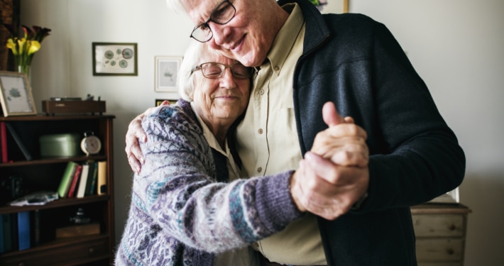 Elderly couple dancing together in a cozy living room with framed artwork and a bookshelf in the background.