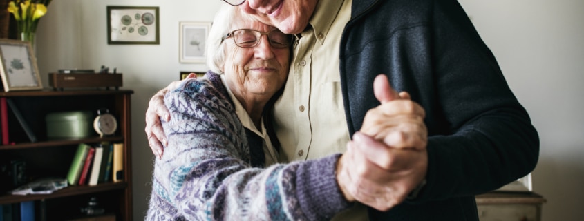 Elderly couple dancing together in a cozy living room with framed artwork and a bookshelf in the background.