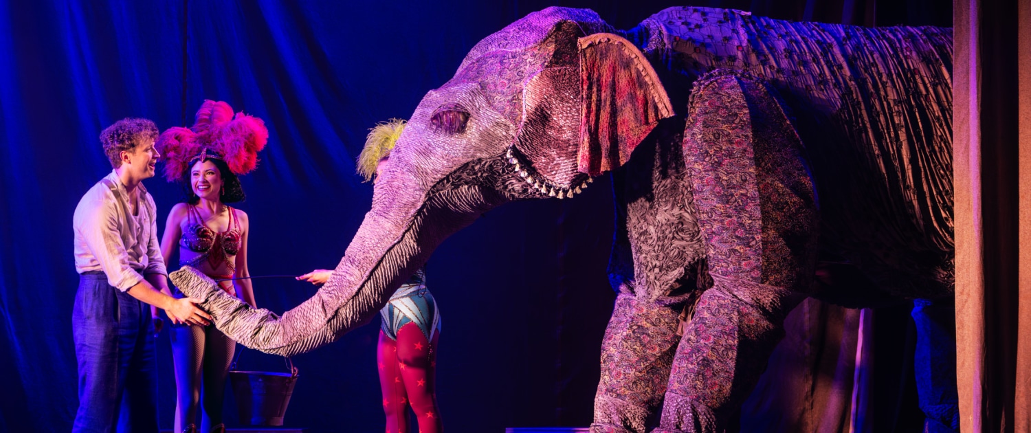 Stage performers interact with a large, elaborate elephant puppet under dramatic theatrical lighting, with colorful costumes and set pieces enhancing the circus-like atmosphere.