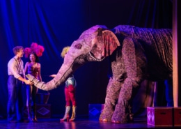 Stage performers interact with a large, elaborate elephant puppet under dramatic theatrical lighting, with colorful costumes and set pieces enhancing the circus-like atmosphere.