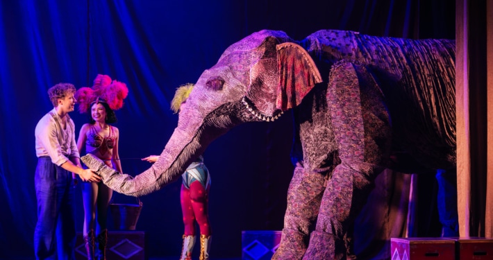 Stage performers interact with a large, elaborate elephant puppet under dramatic theatrical lighting, with colorful costumes and set pieces enhancing the circus-like atmosphere.