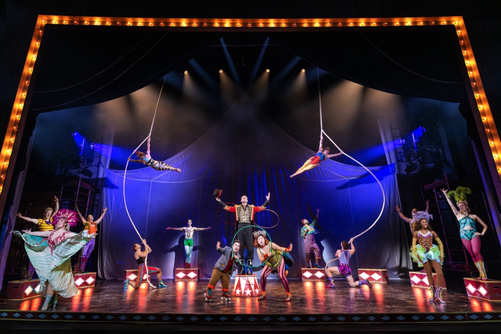 A stage filled with circus acts including arielists, a ring leader, acrobats, and other colorfully dressed performers.