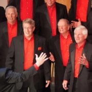 A men’s chorus dressed in black suits with vibrant red shirts singing under the direction of a conductor with arms raised.