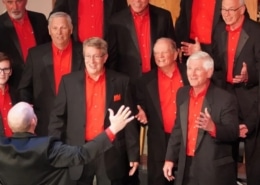 A men’s chorus dressed in black suits with vibrant red shirts singing under the direction of a conductor with arms raised.