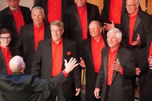 A men’s chorus dressed in black suits with vibrant red shirts singing under the direction of a conductor with arms raised.
