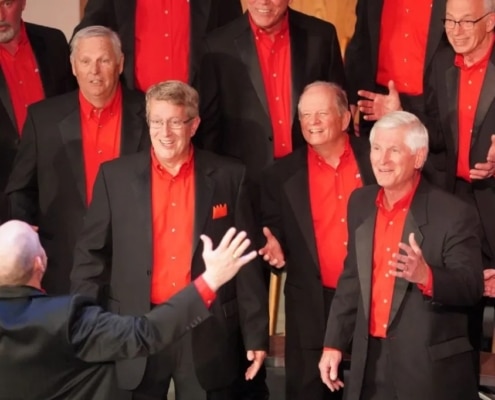 A men’s chorus dressed in black suits with vibrant red shirts singing under the direction of a conductor with arms raised.