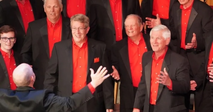 A men’s chorus dressed in black suits with vibrant red shirts singing under the direction of a conductor with arms raised.