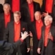A men’s chorus dressed in black suits with vibrant red shirts singing under the direction of a conductor with arms raised.
