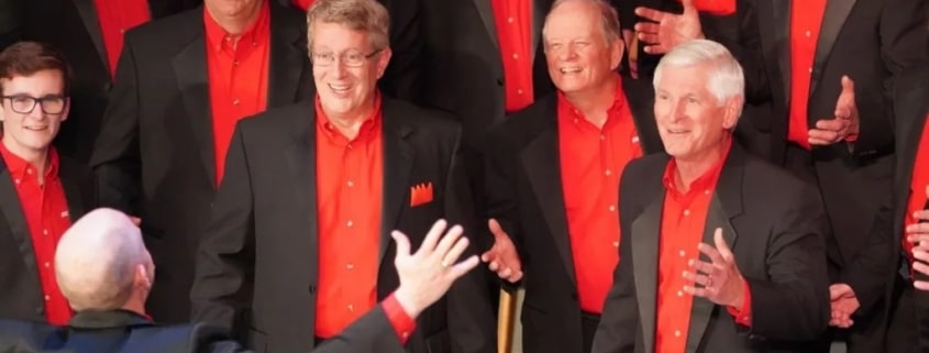 A men’s chorus dressed in black suits with vibrant red shirts singing under the direction of a conductor with arms raised.