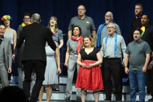 A mixed chorus performing on risers with colorful accessories and costumes, including bright scarves and a red dress, led by a conductor in a dark suit.