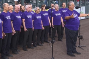 chorus wearing matching purple Colorado Rockies T-shirts singing outdoors near a baseball field with microphones set up in front.