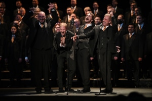 A large choir dressed in black suits with orange ties performing on stage, with several members standing at microphones in the foreground.