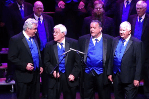 A vocal ensemble wearing black suits with bright blue vests and ties performing on stage, with microphones positioned in front of the group.