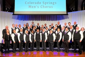 Colorado Springs Men’s Chorus posing on stage in black vests, white shirts, and purple ties under a banner reading “Colorado Springs Men’s Chorus.”