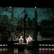 Two performers on a dimly lit stage, one singing and one playing guitar, with a large backdrop depicting a skyline featuring iconic buildings and a reflective water scene.