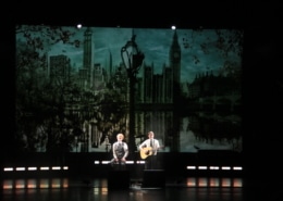 Two performers on a dimly lit stage, one singing and one playing guitar, with a large backdrop depicting a skyline featuring iconic buildings and a reflective water scene.