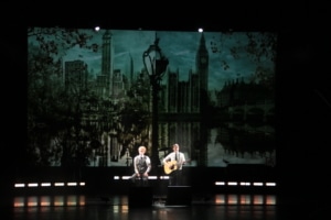 Two performers on a dimly lit stage, one singing and one playing guitar, with a large backdrop depicting a skyline featuring iconic buildings and a reflective water scene.