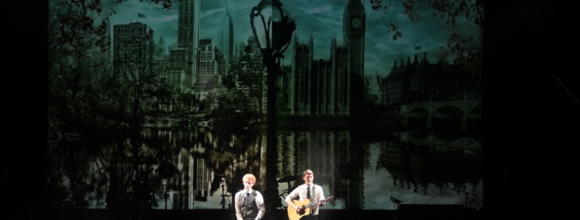 Two performers on a dimly lit stage, one singing and one playing guitar, with a large backdrop depicting a skyline featuring iconic buildings and a reflective water scene.