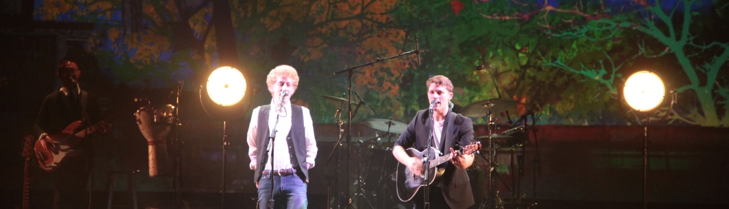 Musicians performing on stage with bright circular lights behind them, one singing at a microphone and the other playing guitar, set against a colorful backdrop with trees and abstract city imagery.