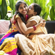 Two individuals wearing traditional colorful attire embracing on a sofa, surrounded by green plants and patterned cushions.