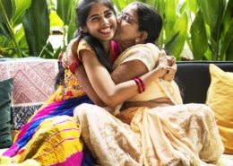Two individuals wearing traditional colorful attire embracing on a sofa, surrounded by green plants and patterned cushions.