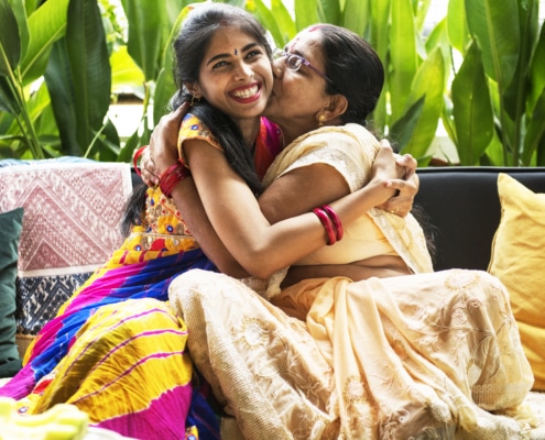 Two individuals wearing traditional colorful attire embracing on a sofa, surrounded by green plants and patterned cushions.