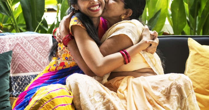 Two individuals wearing traditional colorful attire embracing on a sofa, surrounded by green plants and patterned cushions.