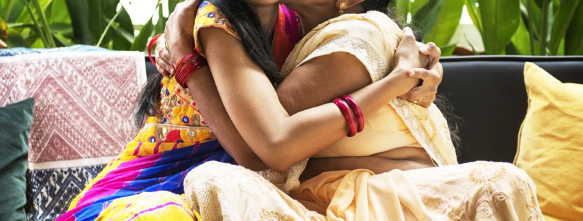 Two individuals wearing traditional colorful attire embracing on a sofa, surrounded by green plants and patterned cushions.