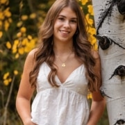 Person with long wavy brown hair wearing a white sleeveless top and jeans, standing outdoors beside a birch tree with yellow autumn foliage in the background.