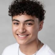 Person with short, dark, curly hair wearing a white shirt and chain necklace, posed against a light gray backdrop.