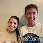 Two people standing side by side indoors against a neutral wall; one wearing a light sweater with an animal graphic, the other wearing a white “Cross Country” T‑shirt and a chain necklace.