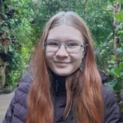 Person with long reddish-brown hair wearing a dark puffer jacket, standing on a wooden pathway surrounded by lush green foliage.